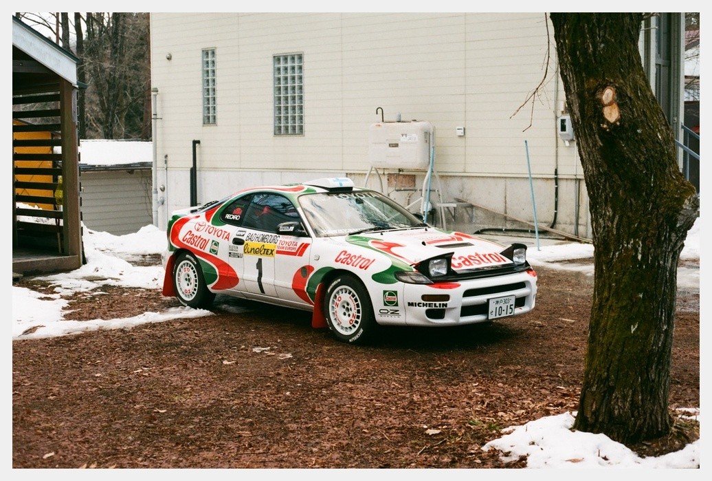 A white rally car with red and green decals is parked on a dirt patch with patches of snow next to a tree and a building.