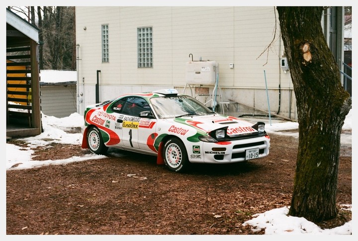 A white rally car with red and green decals is parked on a dirt patch with patches of snow next to a tree and a building.