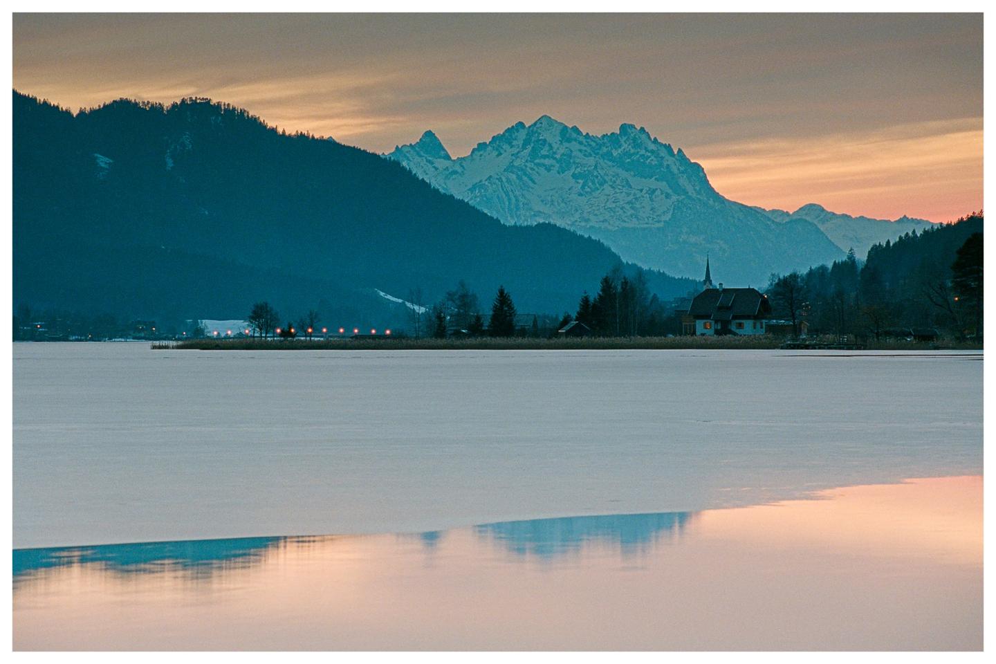 A frozen lake reflects the sunset sky, with mountains and a small village visible in the distance.