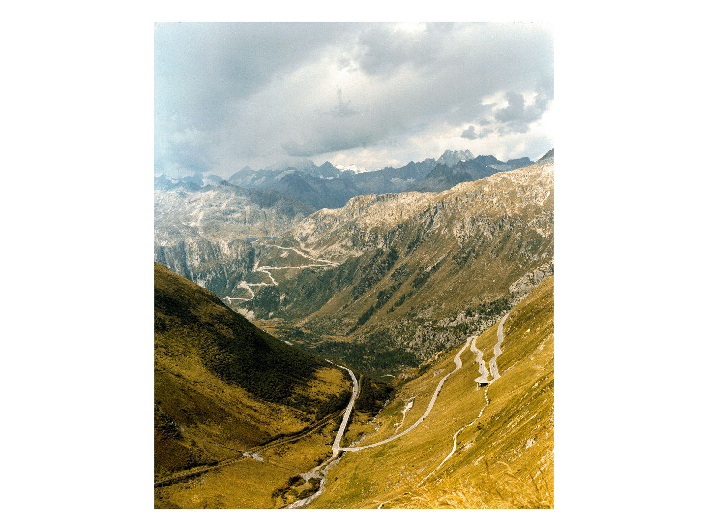 A winding road ascends a steep, grassy mountain valley under a cloudy sky.