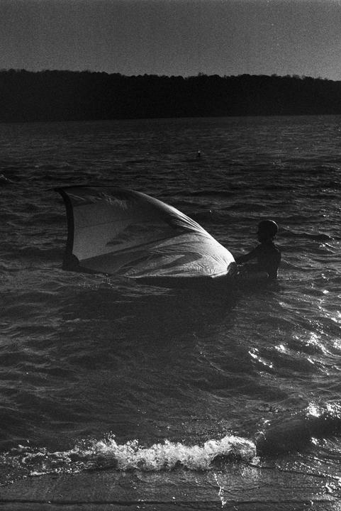A person in a helmet is in the water next to a sail with a dark forest in the background.