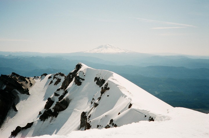 A snow-covered mountain ridge is in the foreground with a larger, snow-capped mountain in the distance under a hazy sky.