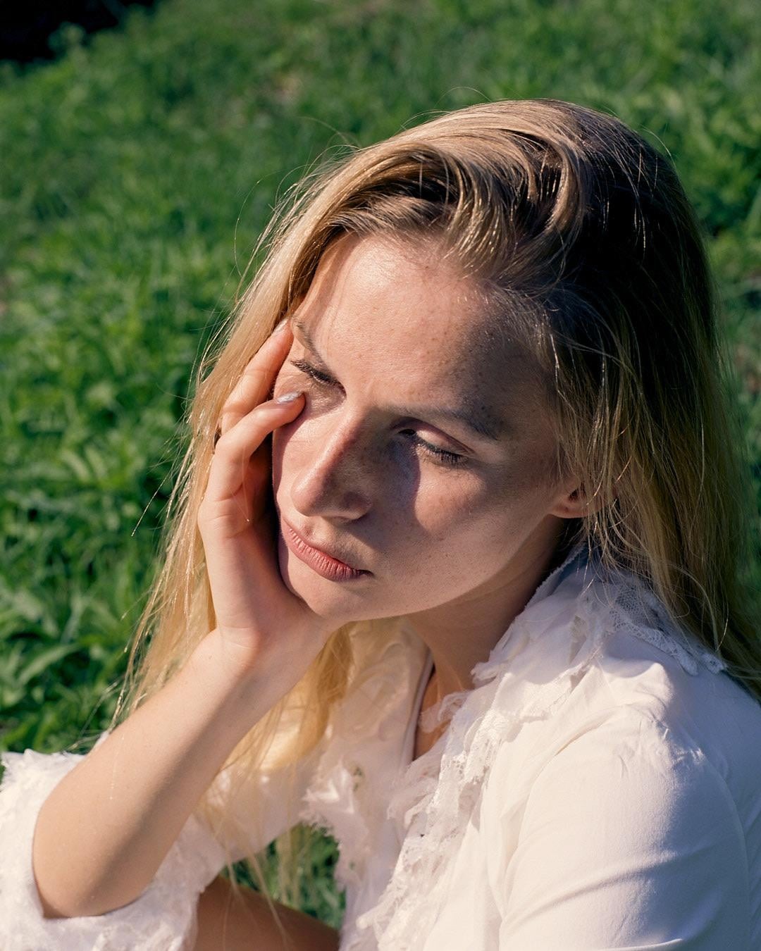 A woman with closed eyes and her hand on her face is sitting in a field of green grass.