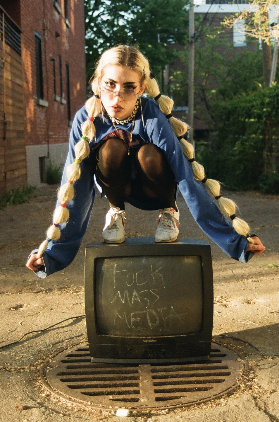 A woman squats on a television set with "FUCK MASS MEDIA" written on its screen, on a street.