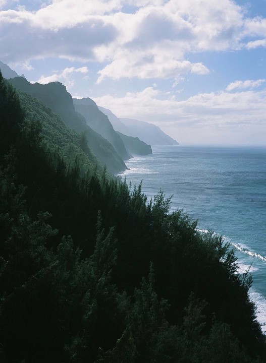 A lush green mountain range meets the ocean under a cloudy sky.