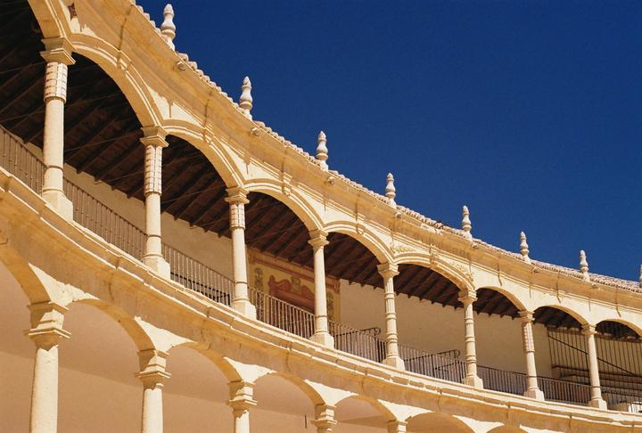 A curved building with multiple arched balconies and columns is shown against a clear blue sky.