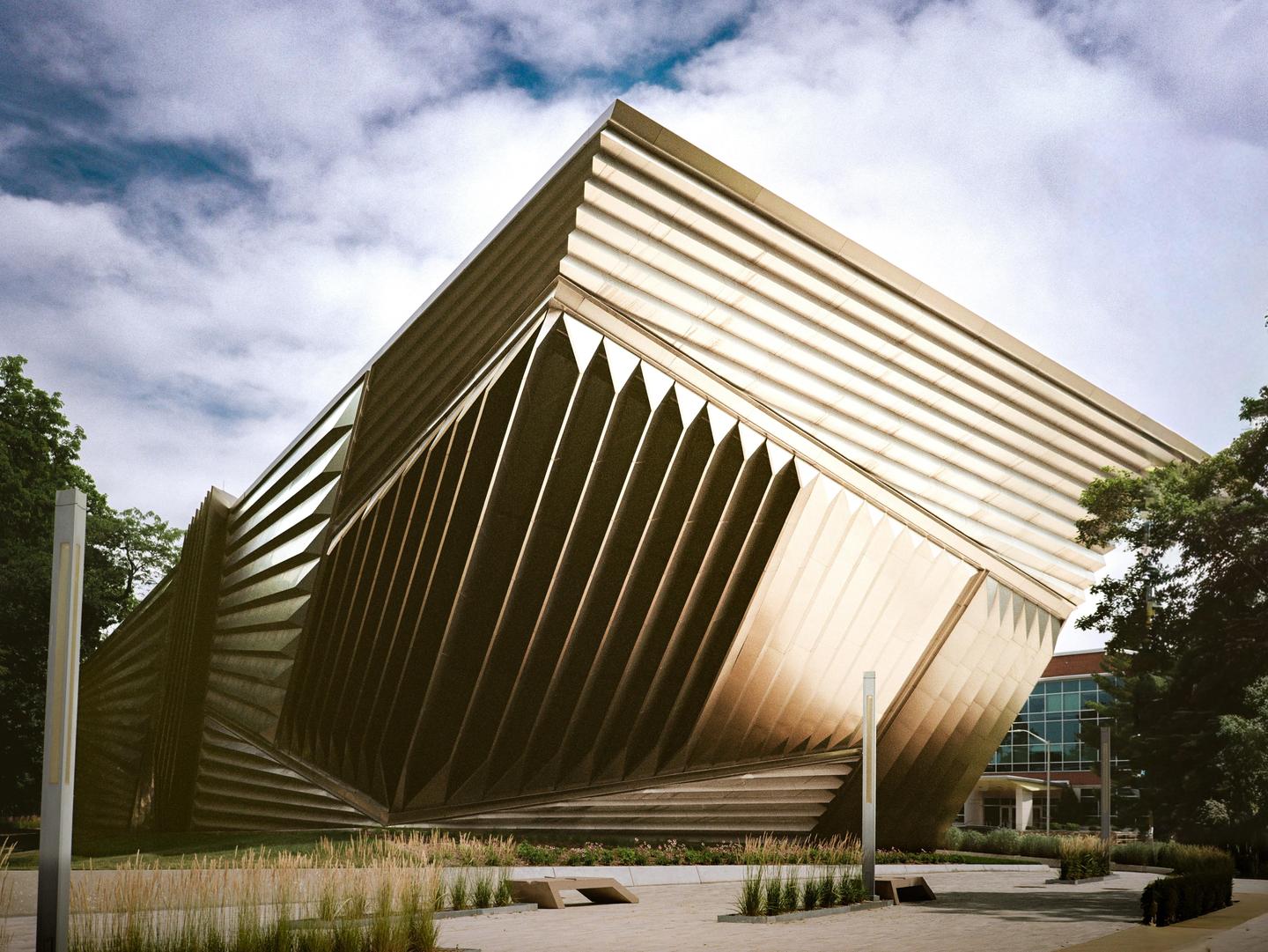 A modern building with a unique geometric facade stands under a cloudy sky, surrounded by trees and landscaping.
