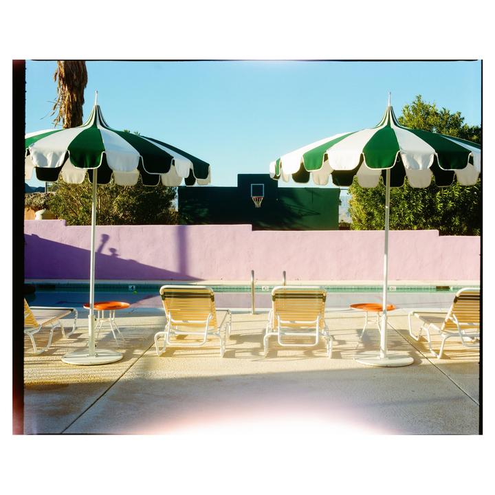 Two green and white striped umbrellas shade lounge chairs next to a swimming pool with a pink wall and a basketball hoop in the background.