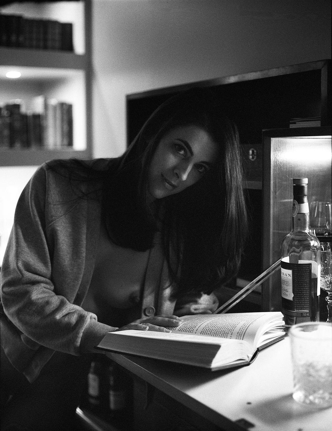 A woman in a cardigan looks at the viewer while leaning over an open book on a counter.