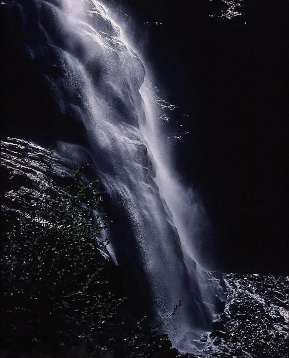 A waterfall cascades down a dark rock face with some foliage visible in the foreground.