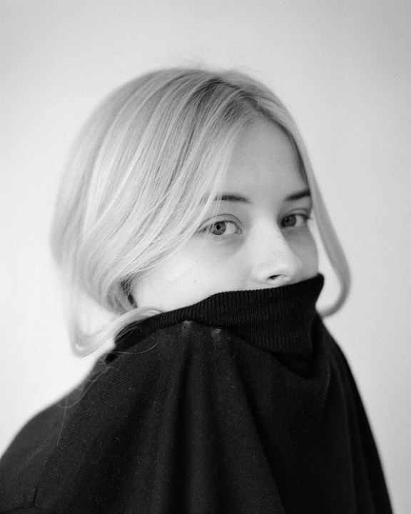 A woman with blonde hair looks over her shoulder, with her face partially covered by a black turtleneck.