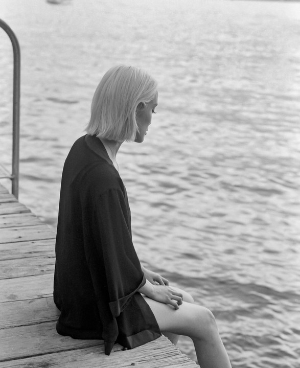 A woman with short blonde hair sits on a wooden dock looking down at the water.