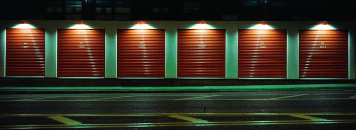Six red garage doors with numbers are illuminated by overhead lights at night, with a street in the foreground.