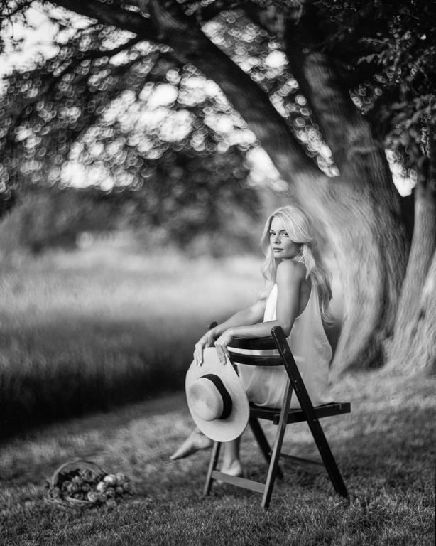 A woman with long hair sits on a chair under a tree, holding a hat, with a basket of flowers nearby.
