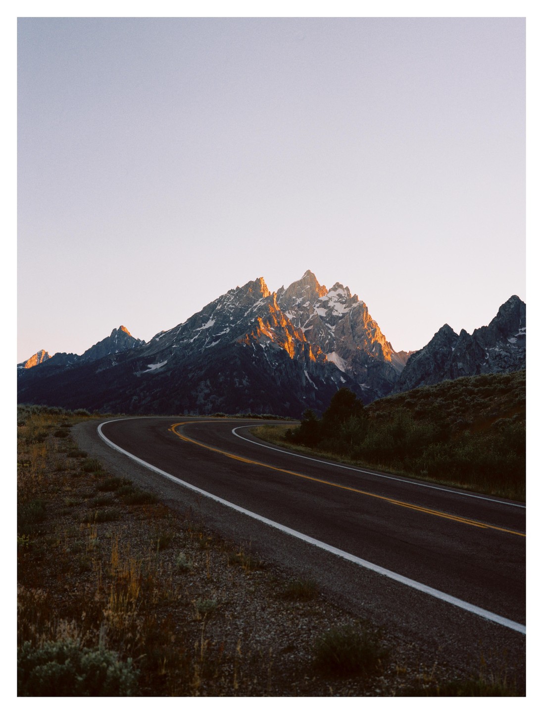 A winding road leads towards a mountain range with sunlit peaks under a clear sky.