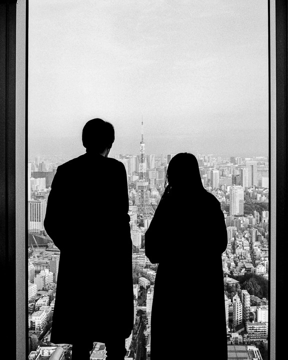 A couple stands silhouetted against a window overlooking the Tokyo cityscape with the Tokyo Tower in the distance.
