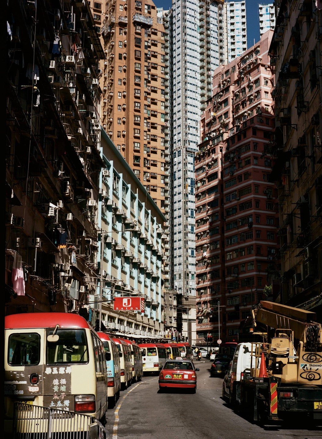 A street in Hong Kong is lined with tall residential buildings and several vehicles, including minibuses and a taxi.