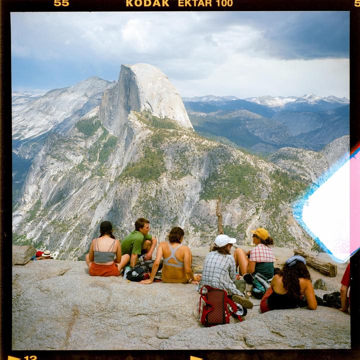 A group of people sit on a rocky outcrop overlooking a vast mountain landscape with a prominent dome-shaped peak.
