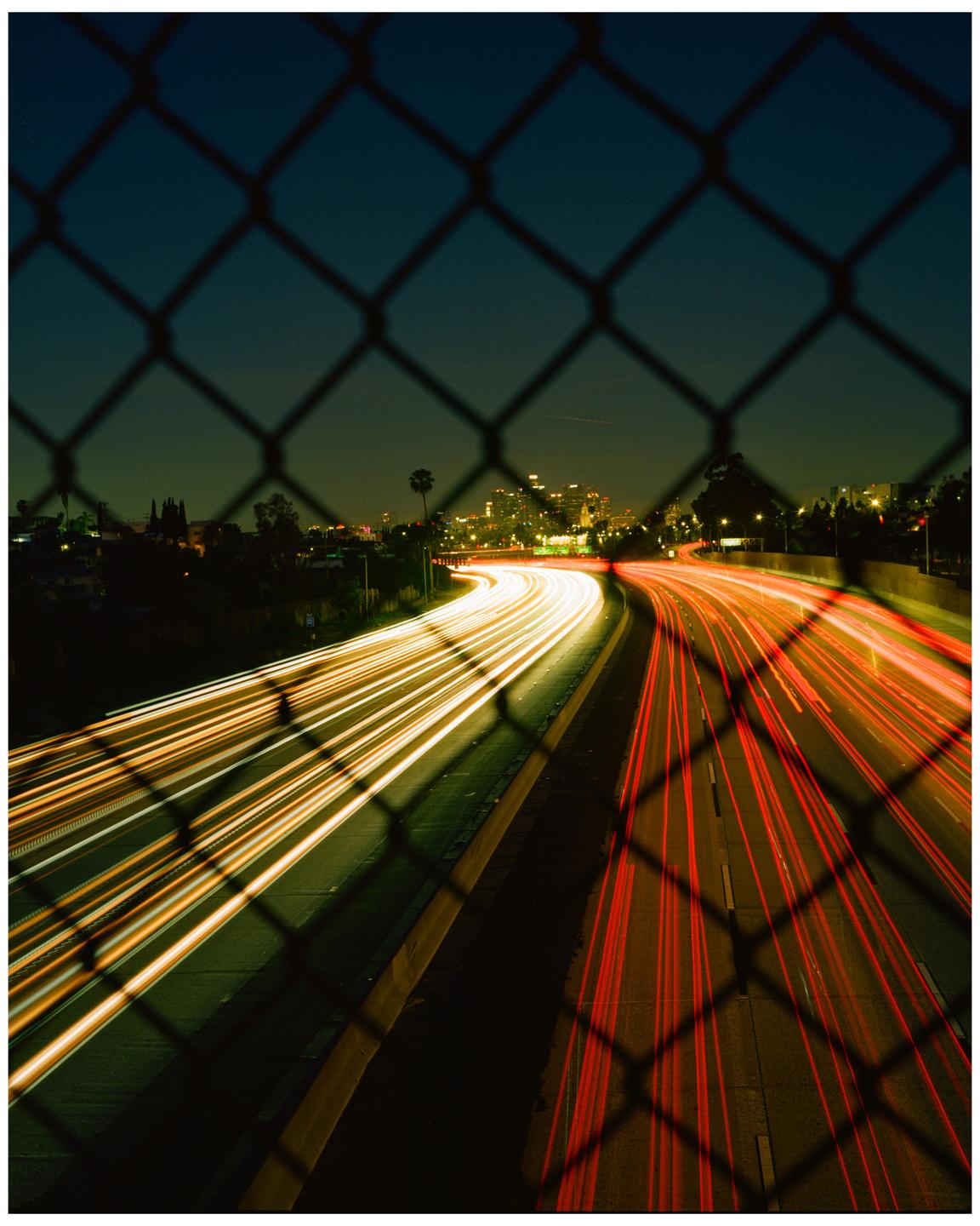 A highway at night is seen through a chain link fence, with light trails from vehicles on the road.