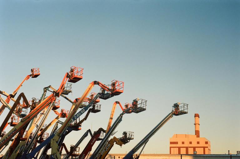 A group of boom lifts are parked in front of a building with a smokestack under a clear sky.