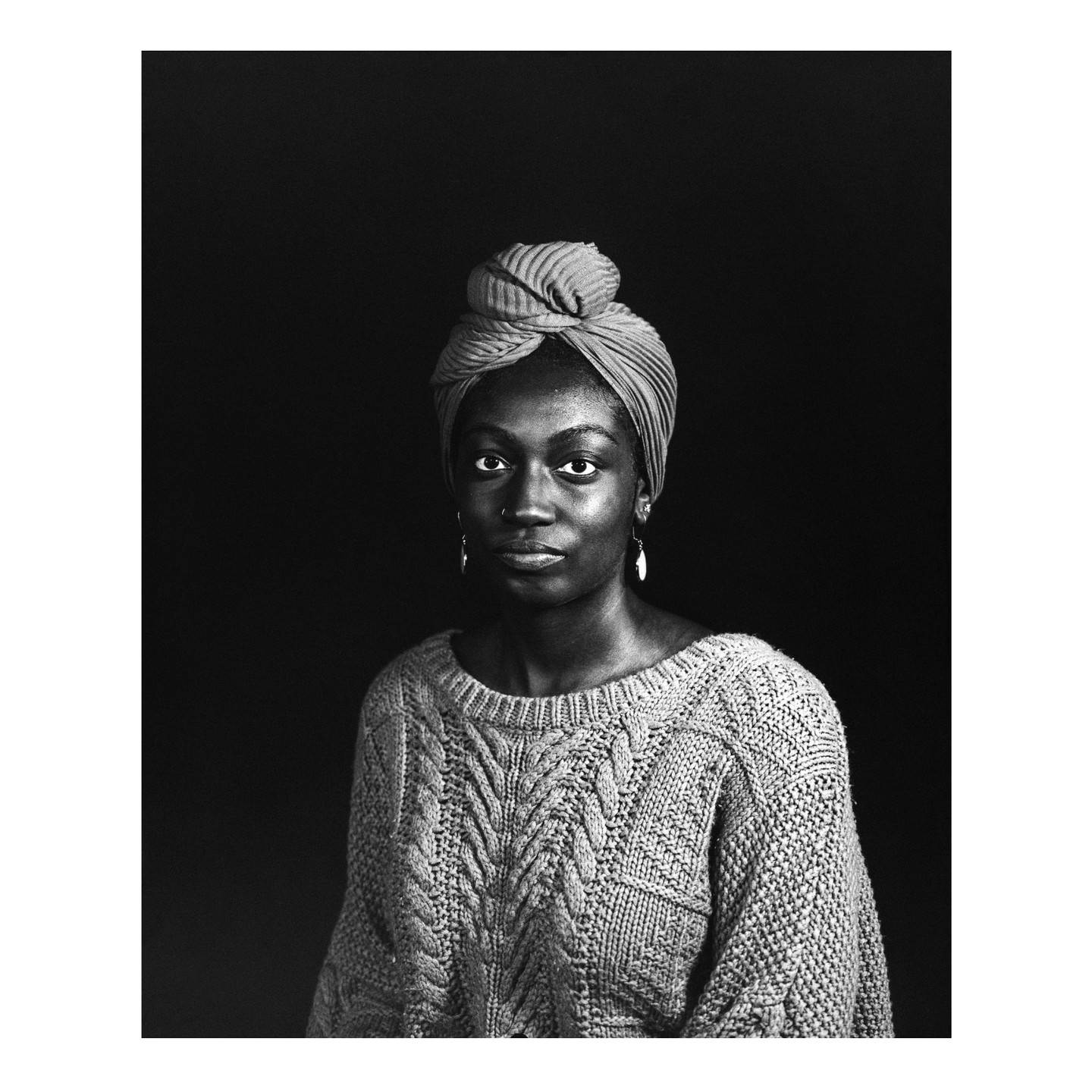 A woman wearing a head wrap and a sweater looks directly at the viewer.