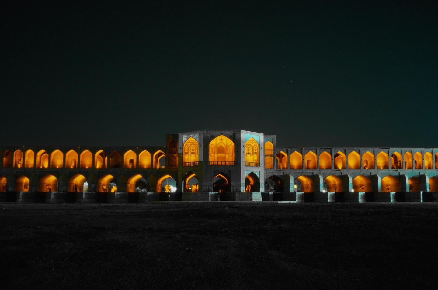 A long, ornate bridge with many arches is illuminated at night, with a dark sky above and dark ground below.