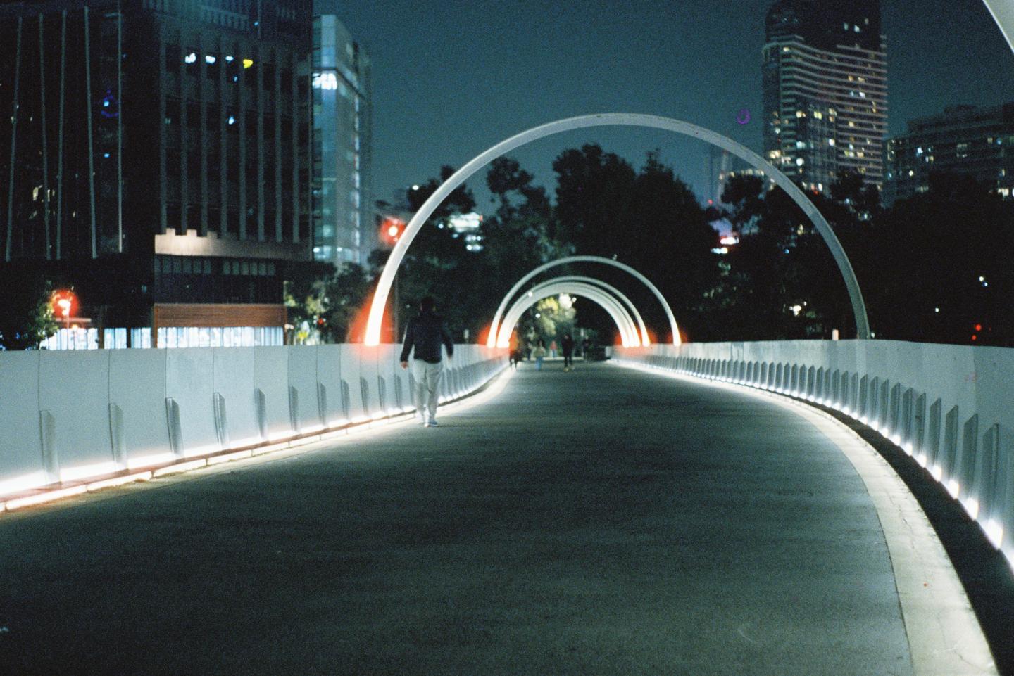 A person walks along a curving pedestrian bridge with illuminated arches and railings at night, with city buildings in the background.