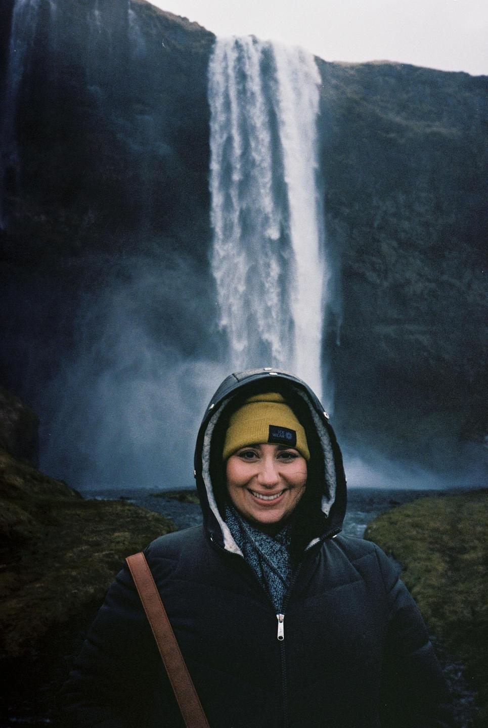 A woman in a winter coat and hat smiles in front of a large waterfall.