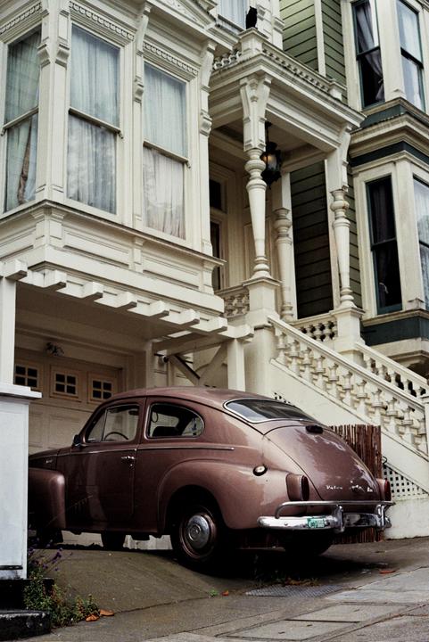 A vintage car is parked in front of a Victorian house with a cat on the porch.