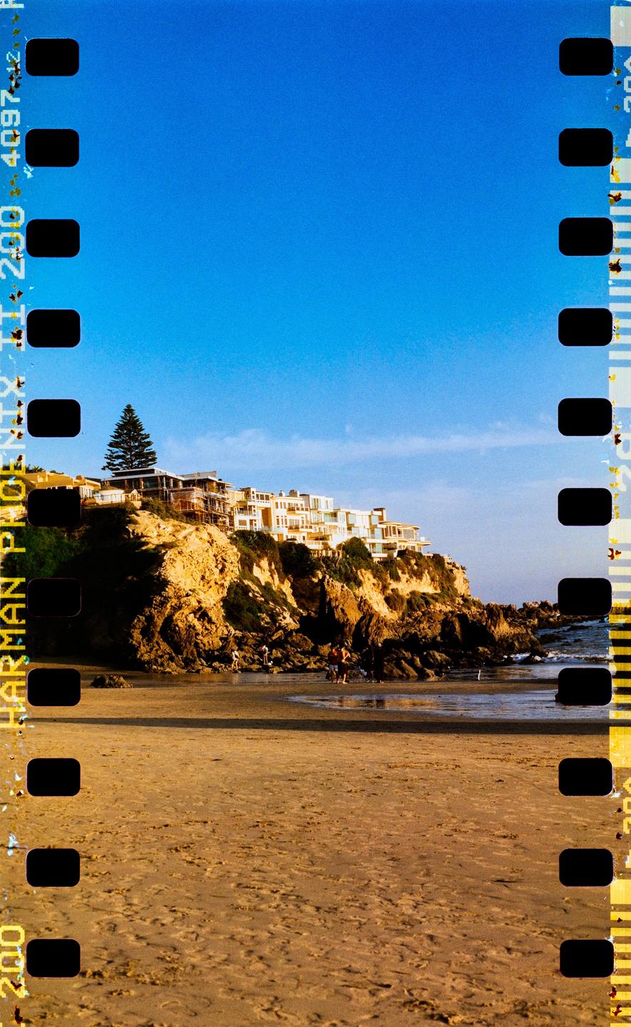 A sandy beach with people walking near the water is overlooked by houses built on a rocky cliff.