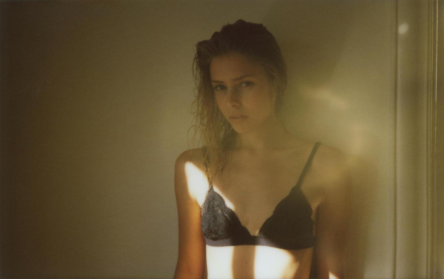 A woman wearing a bra stands in a room with light shining on her body.