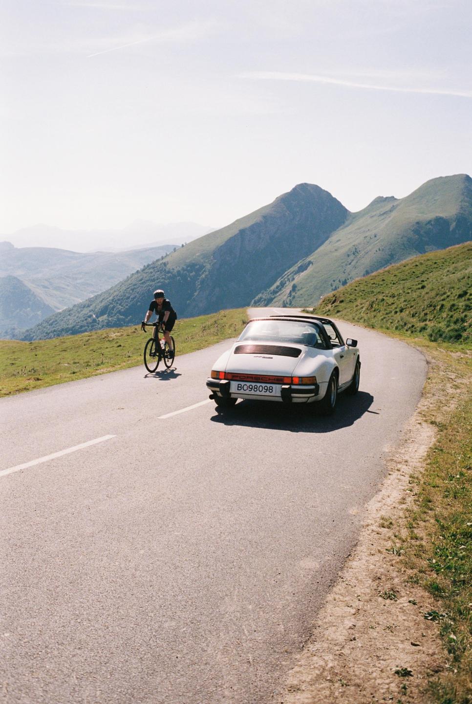 A cyclist rides on a winding mountain road next to a vintage white car.