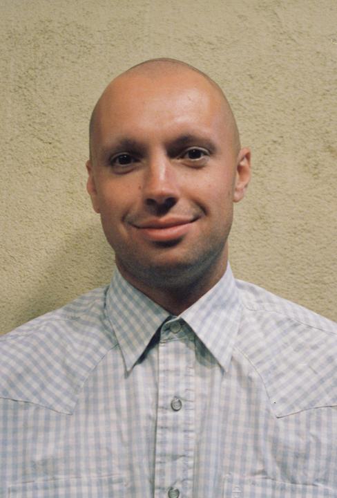 A man with a shaved head and a checkered shirt smiles at the viewer.