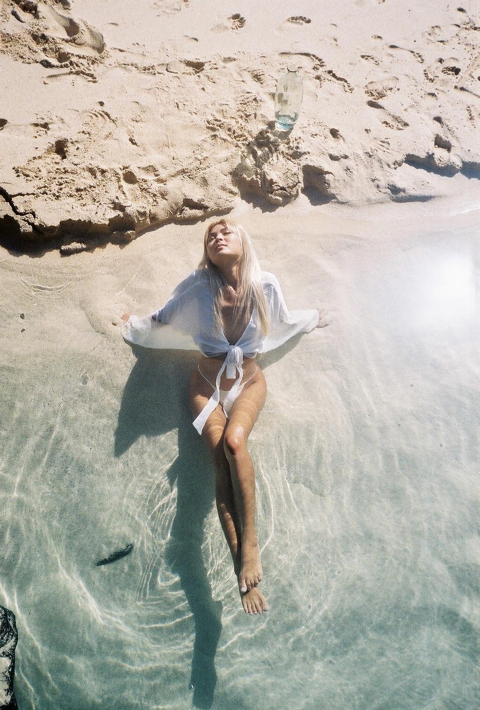 A woman is relaxing in shallow water on a sandy beach with a glass bottle nearby.