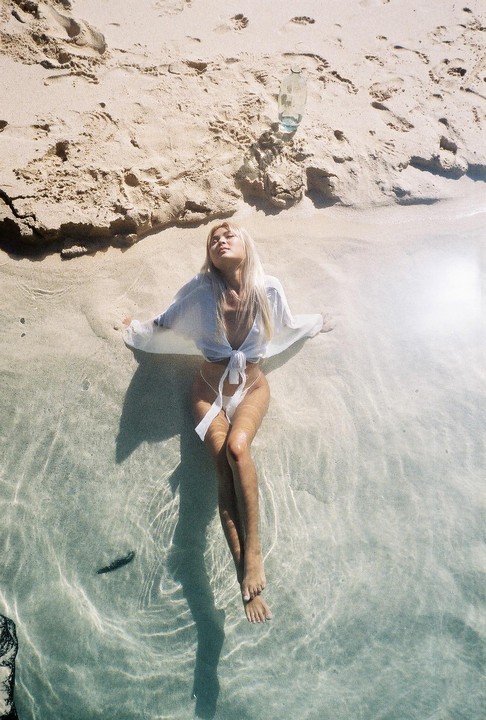 A woman is relaxing in shallow water on a sandy beach with a glass bottle nearby.