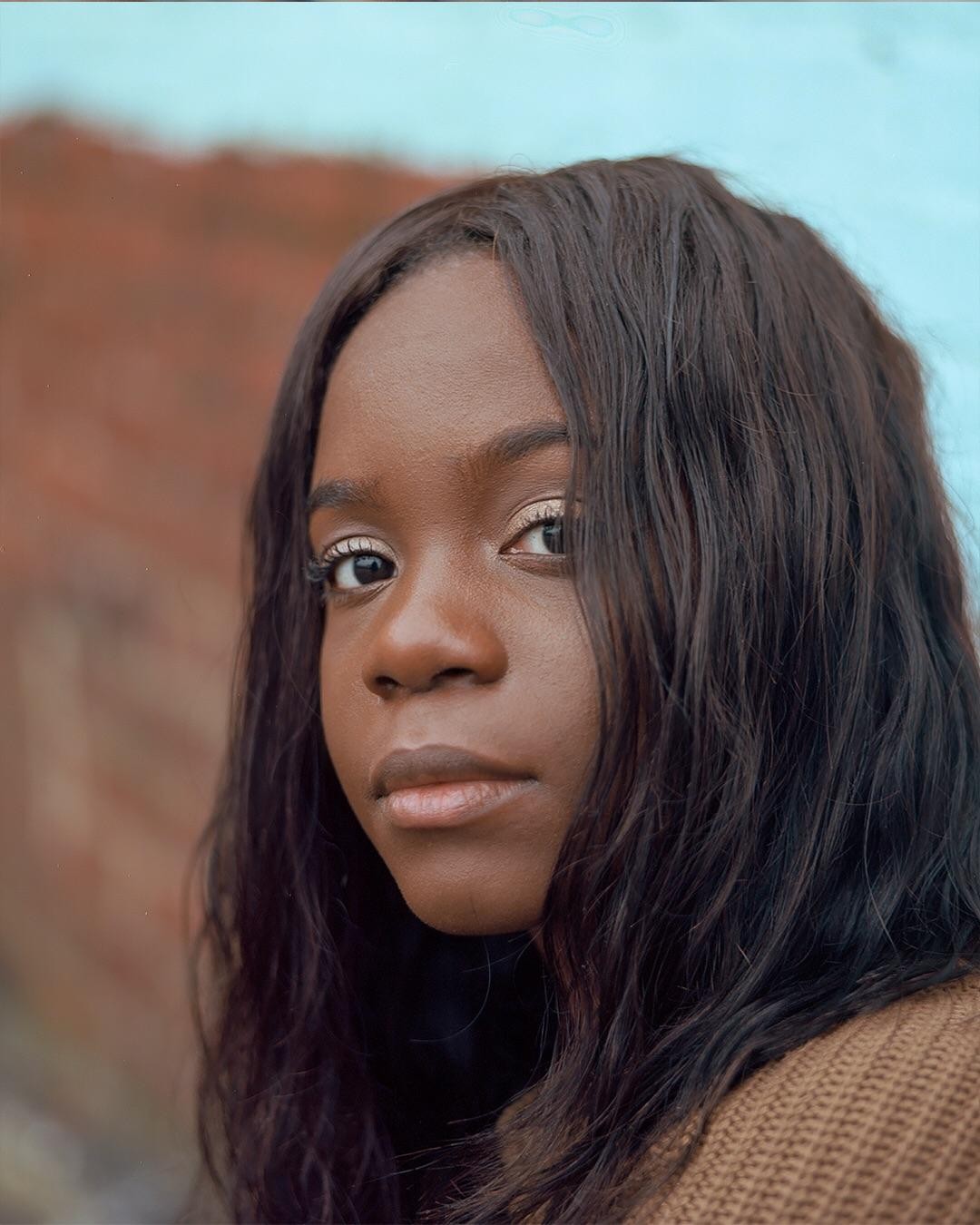 A woman with long dark hair looks directly at the viewer, with a blurred background of a reddish wall and a light blue sky.