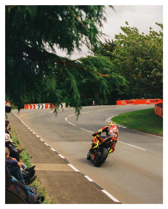 A motorcycle racer leans into a turn on a winding road lined with spectators and trees.