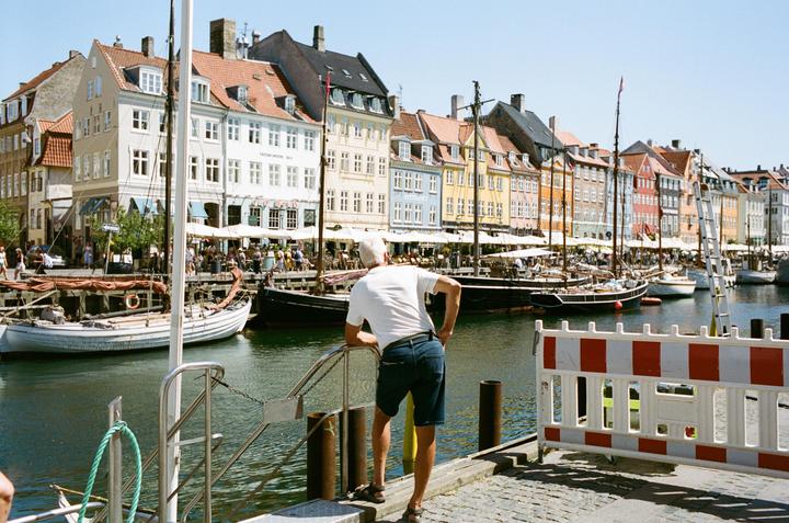 A man stands on a dock looking at boats and colorful buildings across a canal.