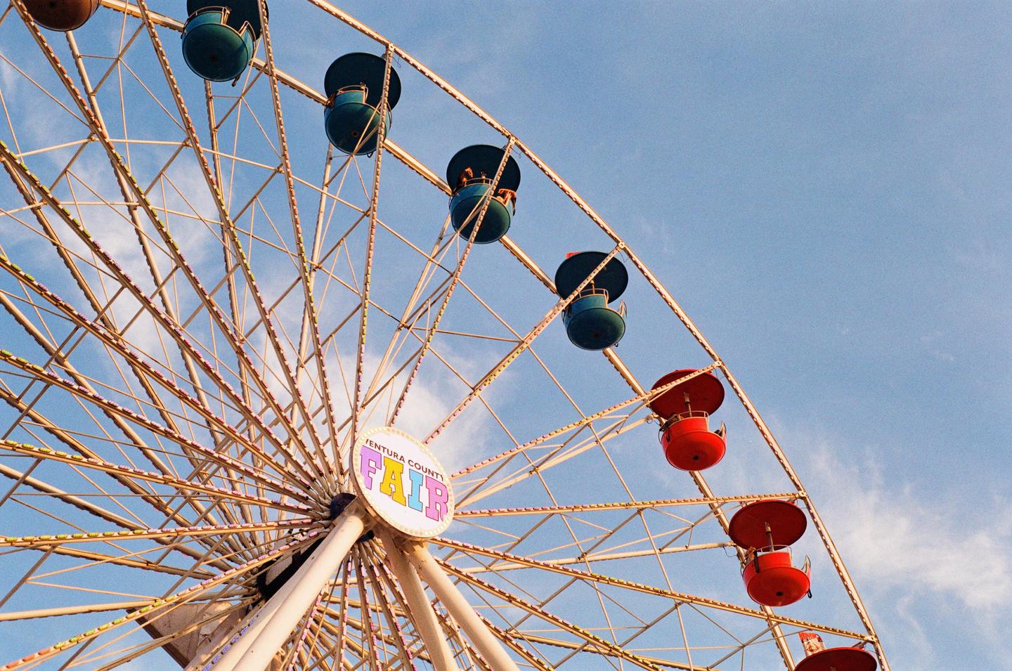 A low-angle view shows a ferris wheel with red and teal gondolas against a blue sky with some clouds.