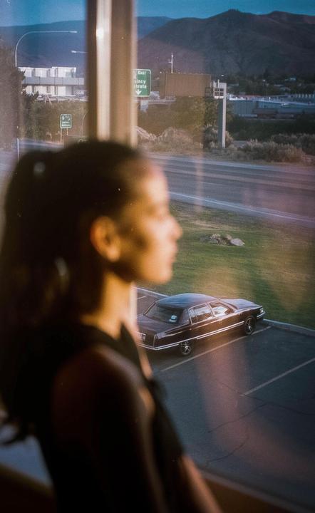 A woman looks out a window at a parking lot with a car, a road, and mountains in the distance.