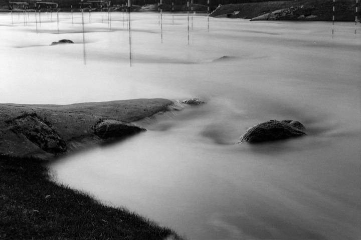 A long exposure shot shows rocks and grass along the edge of a body of water with many poles sticking out of the water in the background.