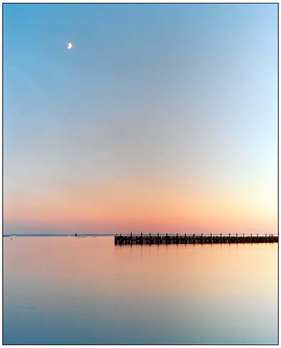 A crescent moon hangs in the sky above a pier stretching into calm water at dusk.