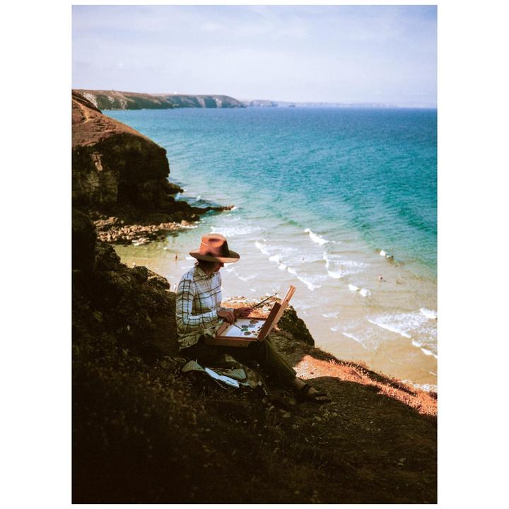 A person wearing a hat is painting on a cliff overlooking a beach and the ocean.