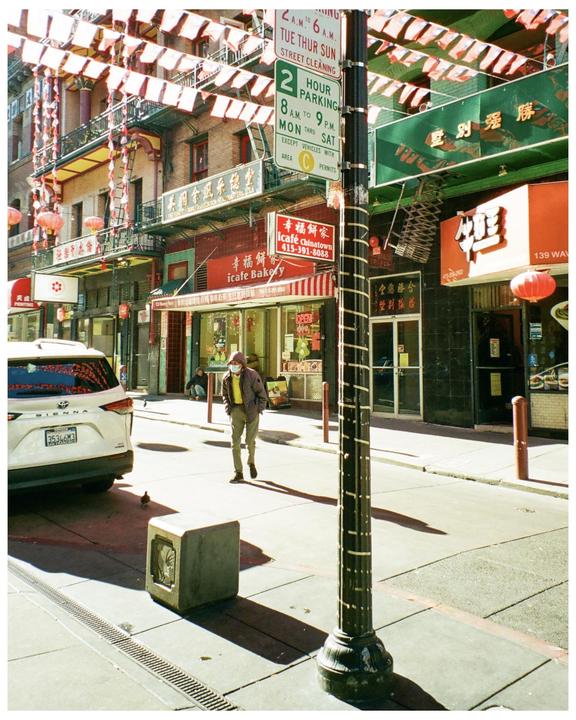 A man walks on a sunny street in Chinatown, San Francisco, with buildings adorned with red lanterns and banners.