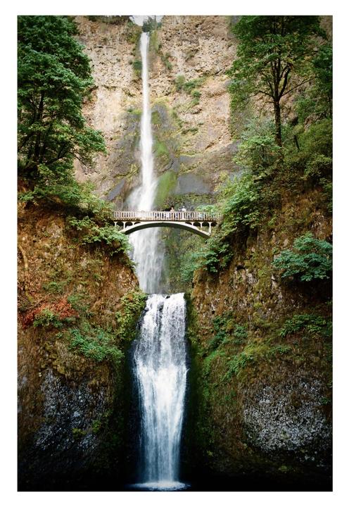 A tall waterfall cascades down a rocky cliff, with a bridge spanning the gorge in front of the upper falls.