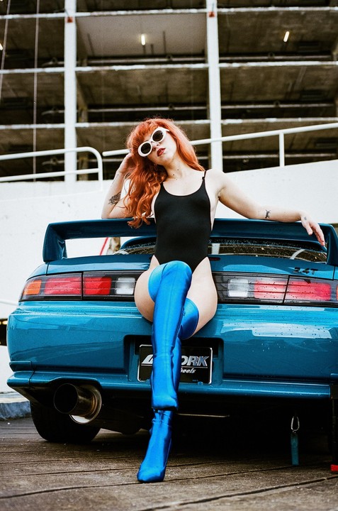 A woman with red hair and sunglasses sits on the trunk of a blue car, wearing a black bodysuit and blue thigh-high boots.