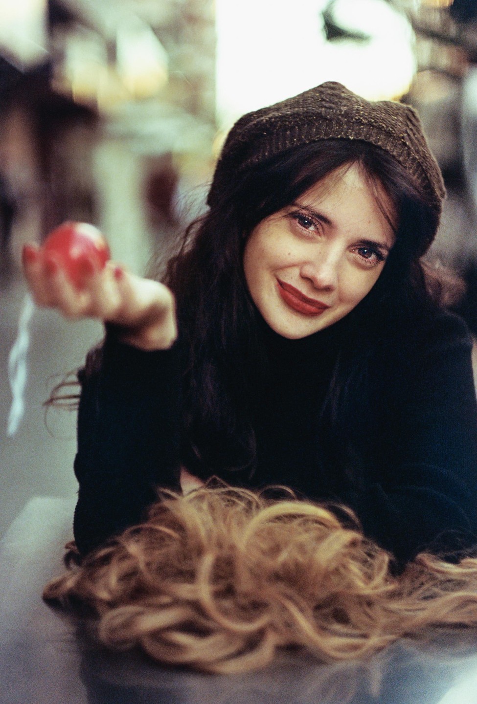 A woman in a dark knit hat and turtleneck smiles at the camera while holding out a red apple.