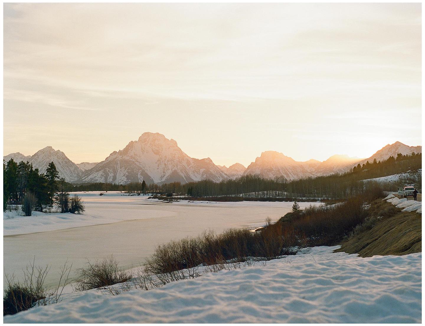A frozen river with snow-covered banks flows past a mountain range under a bright sky.