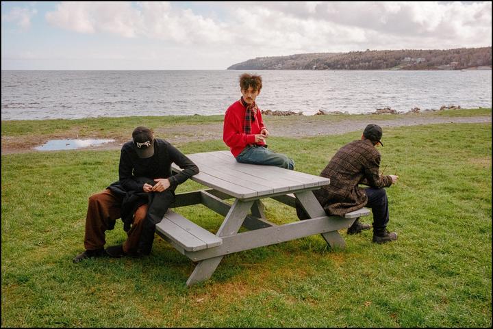 Three men are sitting on and around a picnic table by the water.