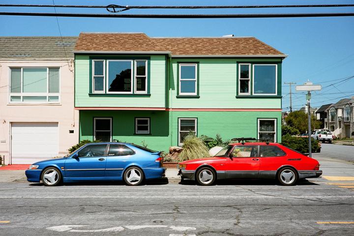 Two cars are parked on a street in front of a green house with a brown roof.